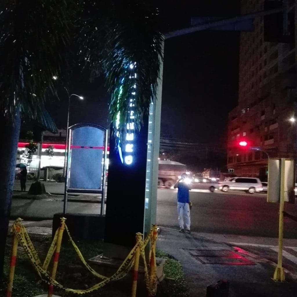 Illuminated pylon signage at night on a Makati street corner, palm tree and barrier tape nearby, by M&G Global Ads.